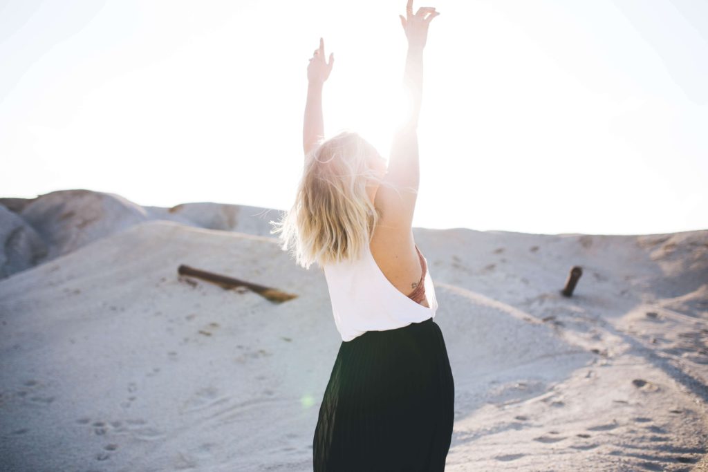 woman stood on sandy dunes in the sun with her arms in the air